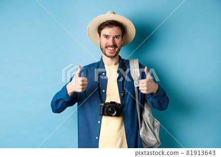 Cheerful handsome man recommending summer vacation place, showing thumbs up and smiling in approval. Tourist leave positive feedback, standing with camera and backpack on blue background Cheerful handsome man recommending summer vacation place, showing thumbs up and smiling in approval. Tourist leave positive feedback, standing with camera and backpack on blue background 81913643