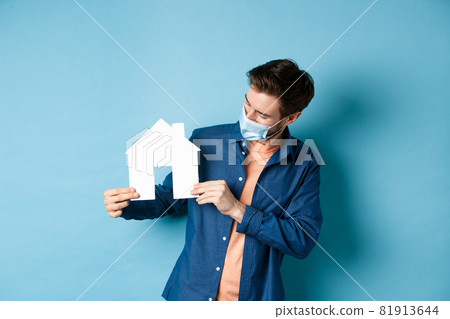 Real estate and quarantine concept. Young caucasian man in medical mask looking at paper house cutout, standing on blue background Real estate and quarantine concept. Young caucasian man in medical mask looking at paper house cutout, standing on blue background 81913644