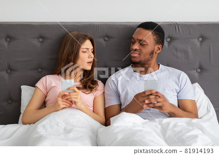 Young interracial couple with smartphones sitting in bed, stuck in gadgets, looking at each other with suspicion Young interracial couple with smartphones sitting in bed, stuck in gadgets, looking at each other with suspicion 81914193