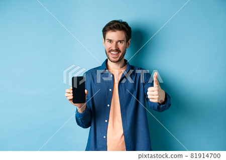 Handsome young man showing thumbs up gesture and empty smartphone screen, recommending app or company, standing on blue background 81914700