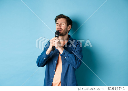 Romantic guy singing in microphone, performing on stage, standing on blue background Romantic guy singing in microphone, performing on stage, standing on blue background 81914741
