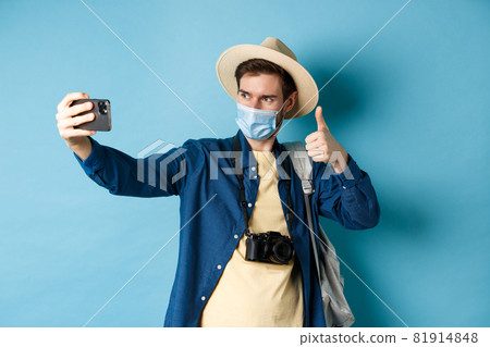 Covid-19, pandemic and travel concept. Happy male tourist taking pictures on vacation, make selfie with thumb up, recommending hotel, standing on blue background Covid-19, pandemic and travel concept. Happy male tourist taking pictures on vacation, make selfie with thumb up, recommending hotel, standing on blue background 81914848
