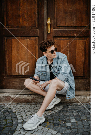 Portrait of happy young man outdoors in front of door, using smartphone. 81914863