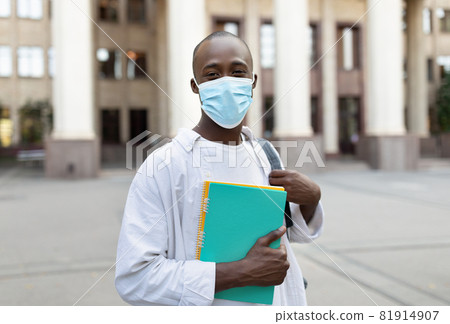 African american male student in face mask standing outdoors with notebooks, posing to camera near university building 81914907