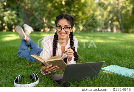 Studentship. Indian student girl learning with laptop and book, taking notes, lying in park outdoors, free space Studentship. Indian student girl learning with laptop and book, taking notes, lying in park outdoors, free space 81914946