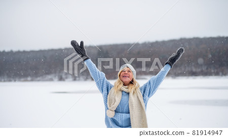 Front view portrait of senior woman outdoors standing in snowy nature, stretching arms. 81914947