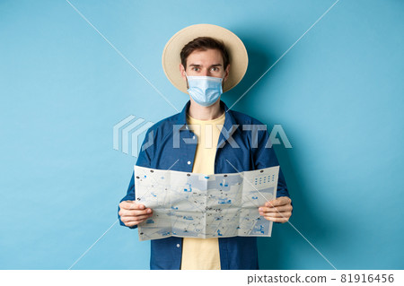 Covid-19, pandemic and travel concept. Happy tourist on summer vacation wearing medical mask and straw hat, holding road map, standing on blue background Covid-19, pandemic and travel concept. Happy tourist on summer vacation wearing medical mask and straw hat, holding road map, standing on blue background 81916456