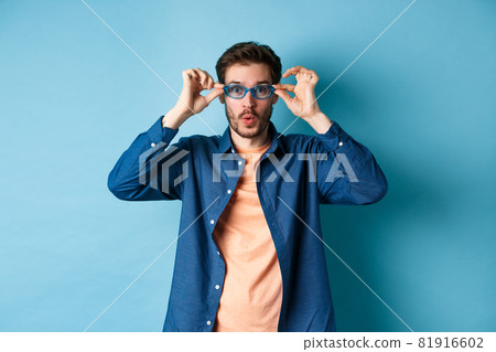 Funny young man trying new sunglasses, looking surprised at camera, standing on blue background Funny young man trying new sunglasses, looking surprised at camera, standing on blue background 81916602