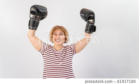 An elderly woman in fighting gloves raises her hands up on a white background. Victory in a duel An elderly woman in fighting gloves raises her hands up on a white background. Victory in a duel 81917180
