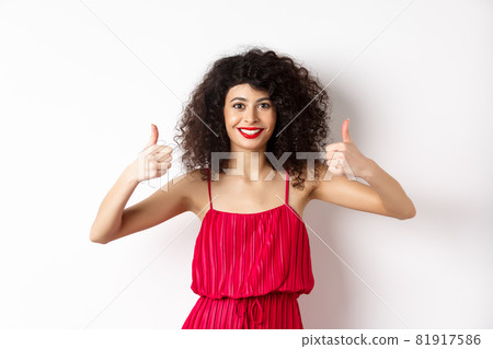Pretty female model with curly hair and red dress, bright makeup, showing thumbs up in approval, say yes, smiling and nodding in approval, white background Pretty female model with curly hair and red dress, bright makeup, showing thumbs up in approval, say yes, smiling and nodding in approval, white background 81917586