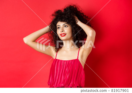 Carefree female model playing with hair, looking dreamy and relaxed at logo, smiling sensual, standing on red background 81917901