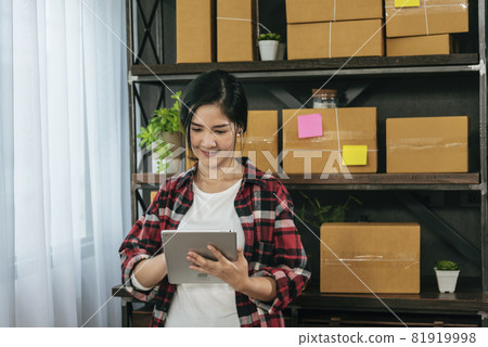 Asian woman startup small business at home office. Online seller entrepreneur young asian woman use tablet checking online order. Woman using tablet check stock goods supply delivery package shipping 81919998