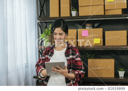Asian woman startup small business at home office. Online seller entrepreneur young asian woman use tablet checking online order. Woman using tablet check stock goods supply delivery package shipping 81919999
