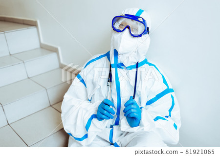 A tired medical worker in a protective jumpsuit and mask sits on the steps of the stairs near the 81921065