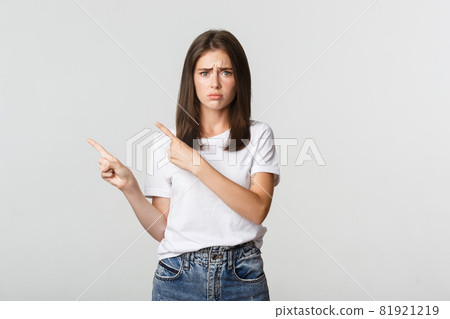 Nervous timid cute girl pointing fingers upper left corner and looking sad, having regret 81921219