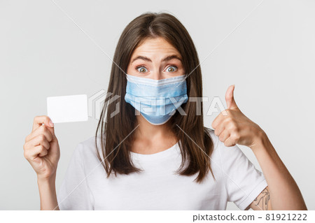 Covid-19, health and social distancing concept. Close-up of pretty brunette girl in medical mask, showing credit card and thumbs-up in approval Covid-19, health and social distancing concept. Close-up of pretty brunette girl in medical mask, showing credit card and thumbs-up in approval 81921222