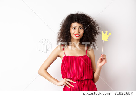 Stylish young woman in red dress, feeling confident and sassy, holding crown and smiling, standing over white background Stylish young woman in red dress, feeling confident and sassy, holding crown and smiling, standing over white background 81922697