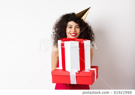 Holidays and celebration. Beautiful lady with curly hair, wearing party hat and holding big presents, smiling happy, receiving birthday gifts, standing on white background 81922998