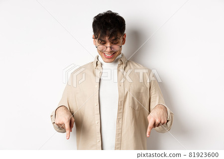 Stylish modern guy in glasses pointing, looking down with happy smile, showing banner, standing on white background Stylish modern guy in glasses pointing, looking down with happy smile, showing banner, standing on white background 81923600