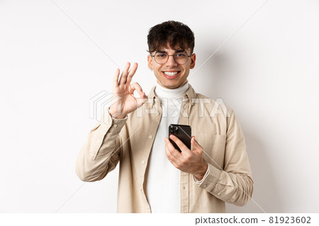 Technology and online shopping concept. Portrait of handsome modern guy in glasses showing OK gesture using smartphone, recommending app or shop, white background Technology and online shopping concept. Portrait of handsome modern guy in glasses showing OK gesture using smartphone, recommending app or shop, white background 81923602