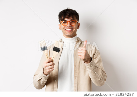 Handsome natural guy in glasses recommending store, showing painting brushes for renovation and decor, make thumbs up in approval, standing on white background Handsome natural guy in glasses recommending store, showing painting brushes for renovation and decor, make thumbs up in approval, standing on white background 81924287