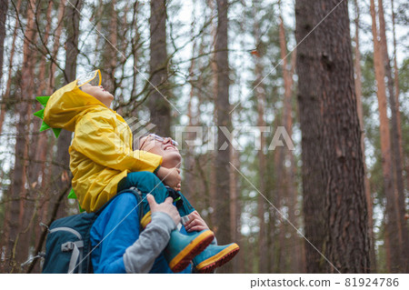 Mom and child walking in the forest after rain in raincoats together, the child is sitting on the shoulders of mom, happily looking at the sky 81924786
