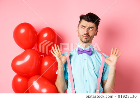 Valentines day concept. Pensive young man in bow-tie, raising hands up and looking left while thinking, making decision, standing on romantic pink background and heart balloons 81925130