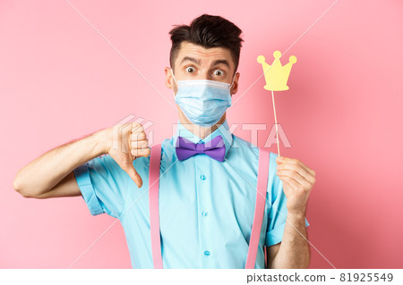 Covid, pandemic and quarantine concept. Disappointed guy in medical mask showing party crown and thumbs down, complaining while standing on pink background 81925549