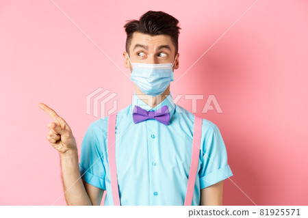 Covid-19, pandemic and health concept. Funny man in bow-tie and medical mask pointing finger left, looking aside curious, showing logo, standing on pink background Covid-19, pandemic and health concept. Funny man in bow-tie and medical mask pointing finger left, looking aside curious, showing logo, standing on pink background 81925571