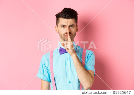 Angry and grumpy man in funny bow-tie shushing at camera, frowning and demand silence, tell to be quiet, scolding someone loud, standing on pink background Angry and grumpy man in funny bow-tie shushing at camera, frowning and demand silence, tell to be quiet, scolding someone loud, standing on pink background 81925647