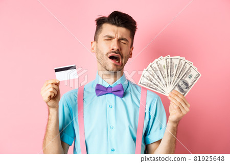 Sad crying man looking miserable, showing plastic credit card and money, standing upset on pink background 81925648
