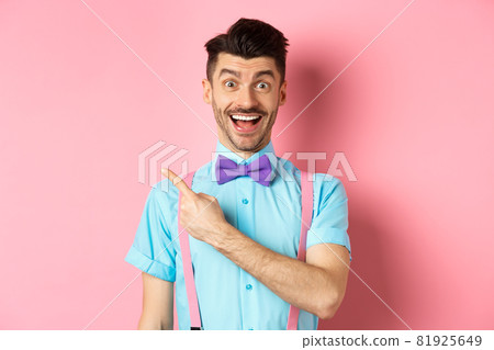 Happy young man checking out promo offer, smiling amazed and pointing finger left at copy space, standing near logo on pink background Happy young man checking out promo offer, smiling amazed and pointing finger left at copy space, standing near logo on pink background 81925649