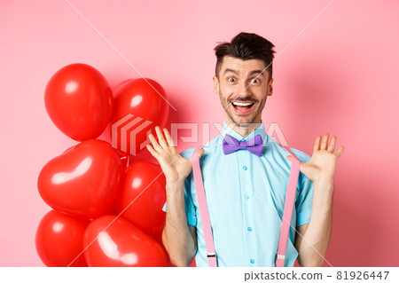 Valentines day concept. Happy young man looking surprised, raising hands up joyful, celebrating near big red hearts and pink background 81926447