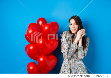Valentines day. Lovely girlfriend ind ress smiling happy at camera, celebrating lovers holiday, standing near heart balloons, blue background 81927387