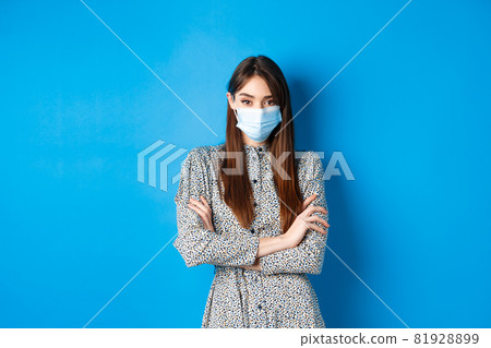 Covid-19, pandemic lifestyle concept. Serious young woman with long natural hair, wearing medical mask, cross arms on chest, protect herself from virus, blue background Covid-19, pandemic lifestyle concept. Serious young woman with long natural hair, wearing medical mask, cross arms on chest, protect herself from virus, blue background 81928899