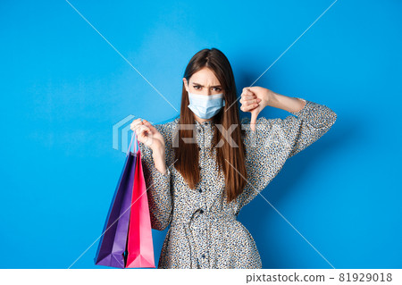 Covid-19, pandemic and lifestyle concept. Angry client in medical mask showing thumb down in dislike, frowning upset, holding shopping bags, blue background 81929018