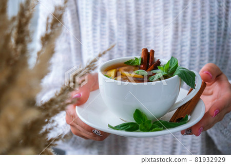 Female hands on sweater holding tea with lemon, cinnamon sticks and mint leaves. Hot drink. Healthy melissa tea natural organic aromatic drink in cup. Autumn winter warming drink. Cozy home 81932929