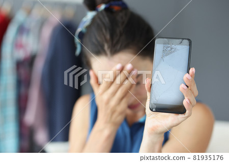 Sad woman covering her eyes with hand and holding broken mobile phone closeup 81935176
