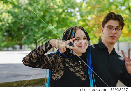A young couple showing victory gestures with their hands against the yellow-green trees. Man and Woman with long blue pigtails. Young adults grimace. Love, relationships. Autumn city park. 81938987