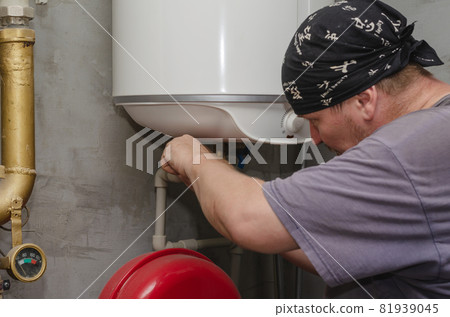 A water heater specialist works in the boiler room. An adult male with a black bandana on his head is installing a wall-mounted water heater. Inside the room. Selective Focus. A water heater specialist works in the boiler room. An adult male with a black bandana on his head is installing a wall-mounted water heater. Inside the room. Selective Focus. 81939045