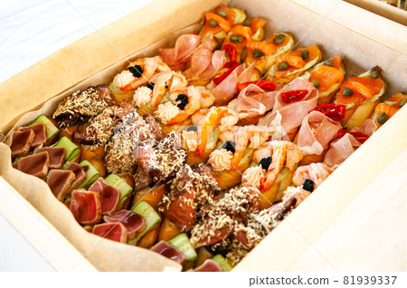 Box with small sandwiches, bruschetta with cold cuts, cheese and seafood. Gourmet snack food in cardboard box for catering buffet for party. Box with small sandwiches, bruschetta with cold cuts, cheese and seafood. Gourmet snack food in cardboard box for catering buffet for party. 81939337