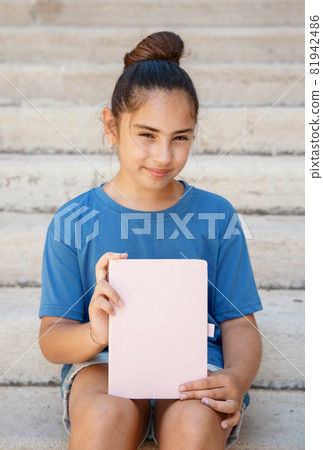 Kid girl in blue t-shirt sitting on a stairs and holding notebook in hands 81942486