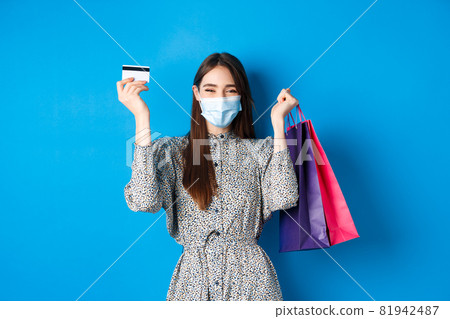 Covid-19, pandemic and lifestyle concept. Pretty female model in medical mask, rejoicing over shopping discounts, holding plastic credit card and bags, standing happy on blue background 81942487