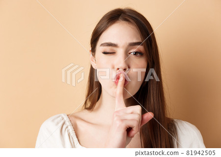 Close up of attractive woman shushing at camera, hiding secret, making hush sign and winking, show taboo sign, standing on beige background 81942505