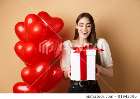 Happy woman open Valentines day gift, smiling excited and looking at present box, standing near red heart balloons on beige background Happy woman open Valentines day gift, smiling excited and looking at present box, standing near red heart balloons on beige background 81942590