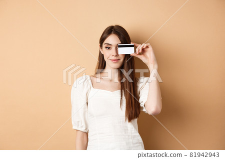 Smiling pretty girl showing plastic credit card over eye and looking satisfied, standing on beige background. Concept of shopping Smiling pretty girl showing plastic credit card over eye and looking satisfied, standing on beige background. Concept of shopping 81942943