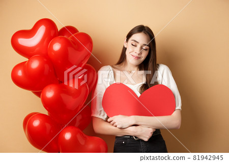 Romantic woman hugging big red heart and smiling dreamy, falling in love on Valentines day, dreaming about lover, standing on beige background near balloons Romantic woman hugging big red heart and smiling dreamy, falling in love on Valentines day, dreaming about lover, standing on beige background near balloons 81942945