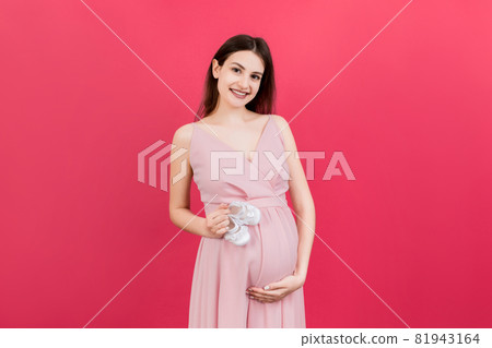 Happy pregnant woman holding baby girl booties against her belly at Colored background. Happy pregnancy. Copy space 81943164