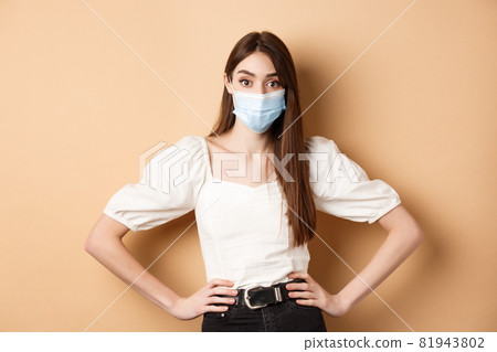 Covid-19 and lifestyle concept. Woman in medical mask looking alarmed at camera, standing on beige background 81943802