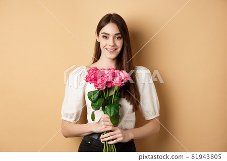 Romantic Valentines day concept. Beautiful young lady holding pink roses and smiling, standing happy on beige background 81943805